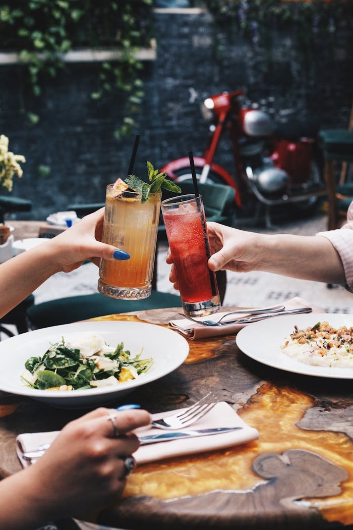 Two people clinking cocktails over a meal in an outdoor café setting, capturing a moment of enjoyment.