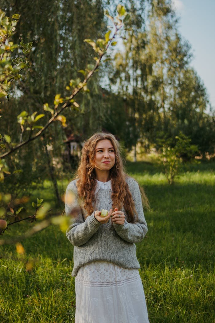 A woman in a lush orchard, holding a green apple, amidst sunny greenery.