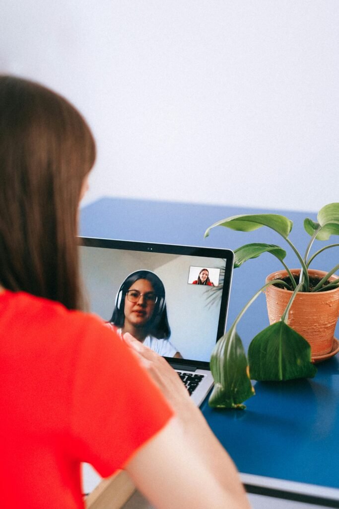 Woman in red shirt video chatting on laptop with plant nearby at home.